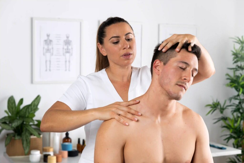 A female therapist in a white tunic giving a neck and shoulder massage in a bright spa room with plants and skincare products